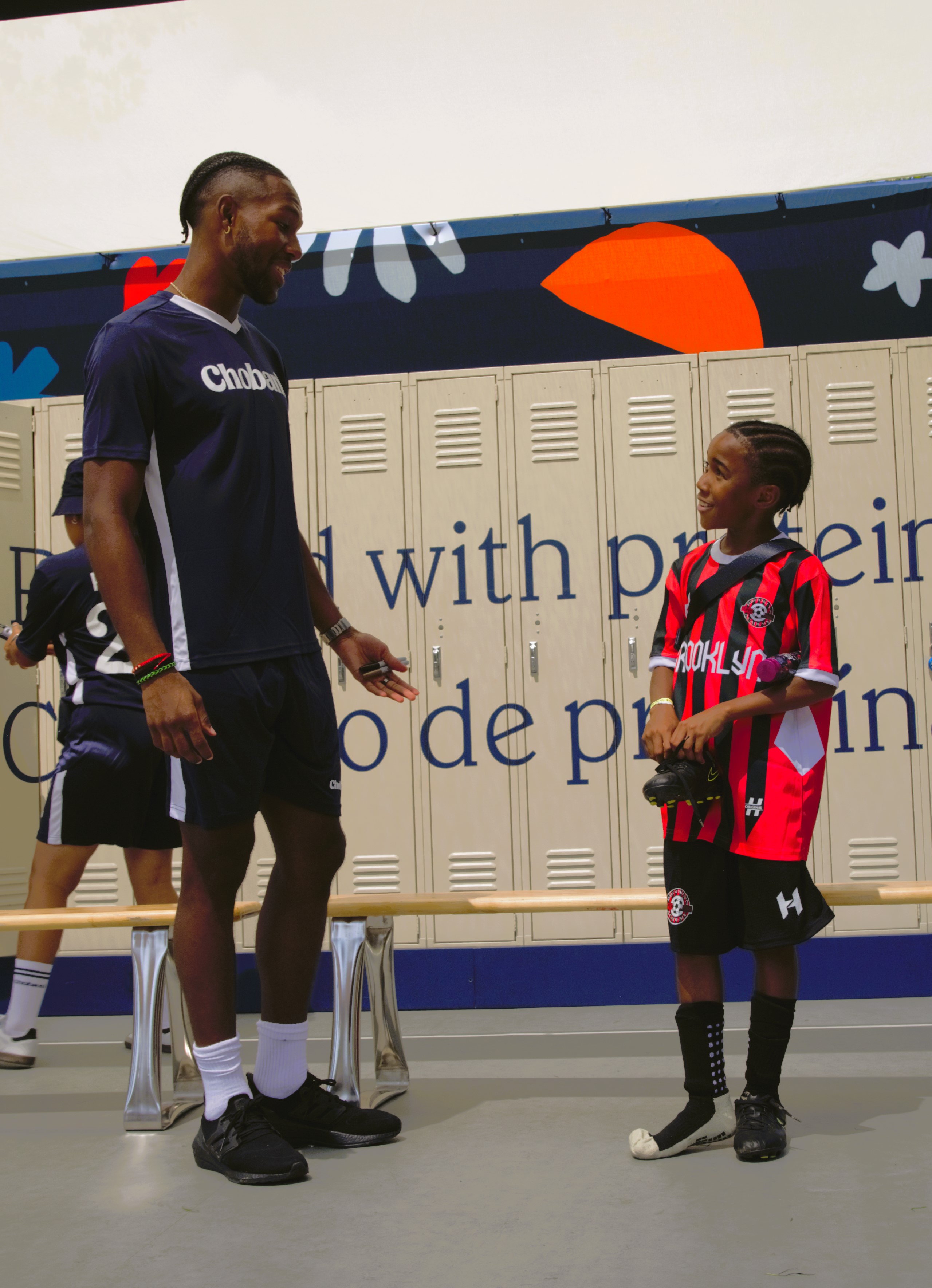 Andres Perea in a Chobani kit, smiling down at a boy in a Brooklyn jersey.